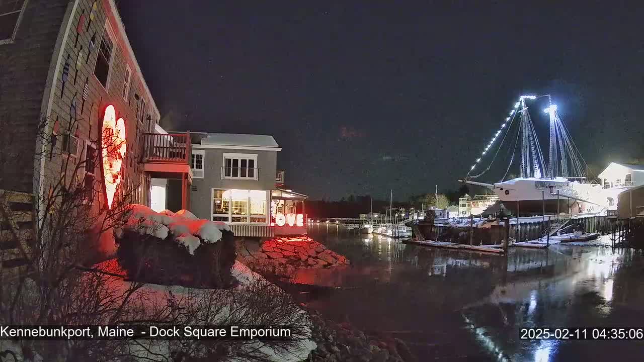 A nighttime scene of a snow-covered waterfront with a large illuminated ship and buildings decorated with red heart and white LOVE lights.