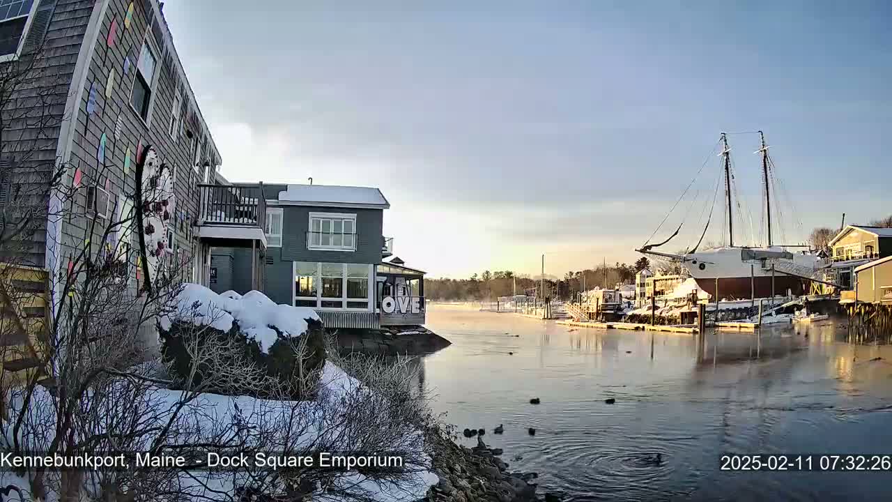 A snow-covered waterfront scene shows a calm body of water with a large sailboat docked, and several buildings nearby under a clear, bright sky.