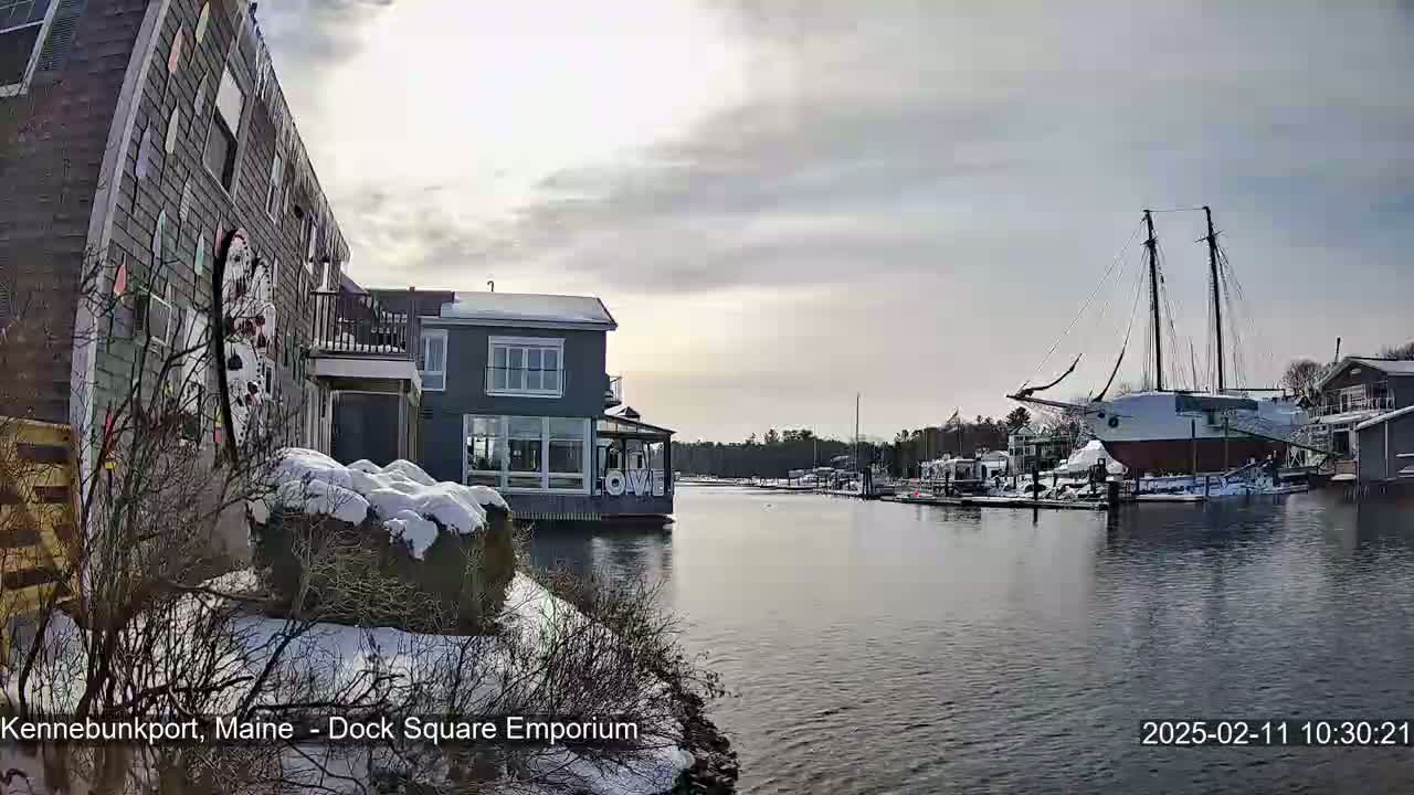 A snow-covered waterfront scene shows a large sailboat docked near buildings, with a calm body of water in the foreground under a partly cloudy sky.