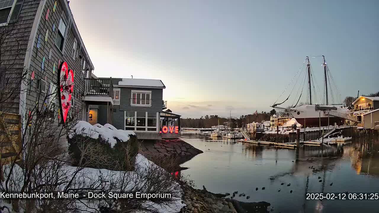 A snow-covered waterfront scene at dusk shows a building with a large heart-shaped light display next to a calm waterway with a docked sailboat and other buildings.