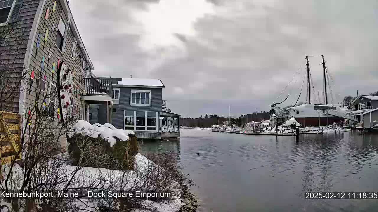 A snow-covered waterfront scene shows buildings, a docked sailboat, and calm water under an overcast sky.