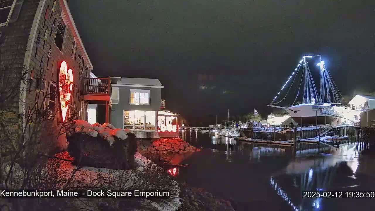 A nighttime scene of a snow-covered waterfront shows buildings lit up, a large sailboat docked, and a red heart-shaped light on a building.
