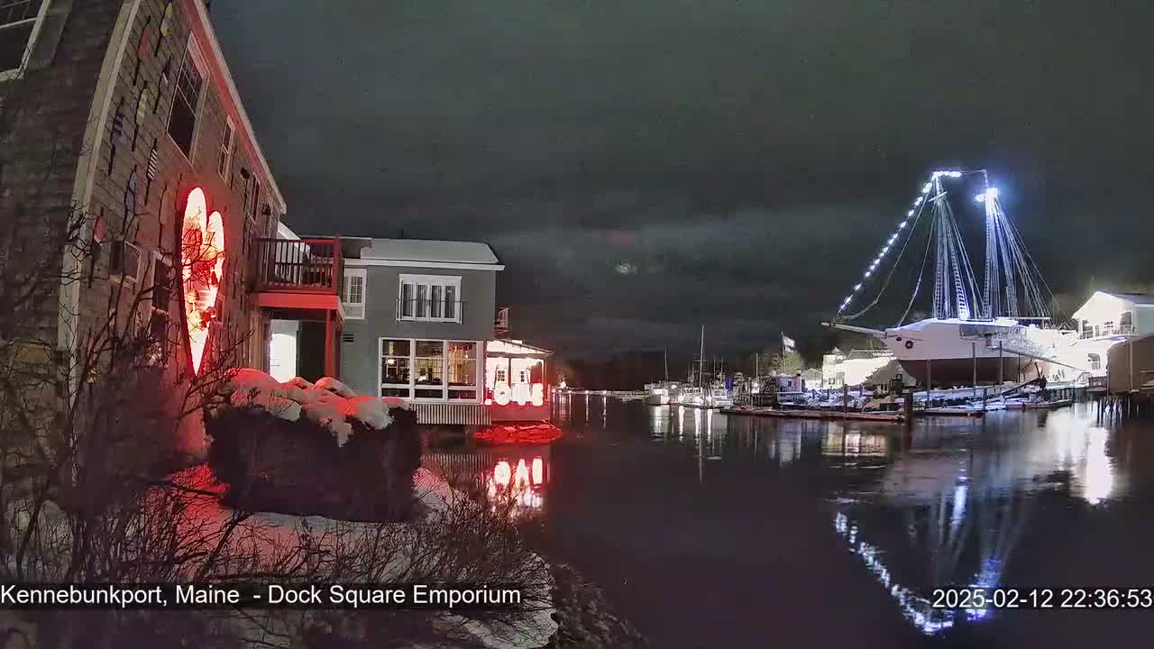 A large illuminated heart is displayed on a building beside a snow-covered waterfront at night, with a large illuminated sailboat docked nearby.