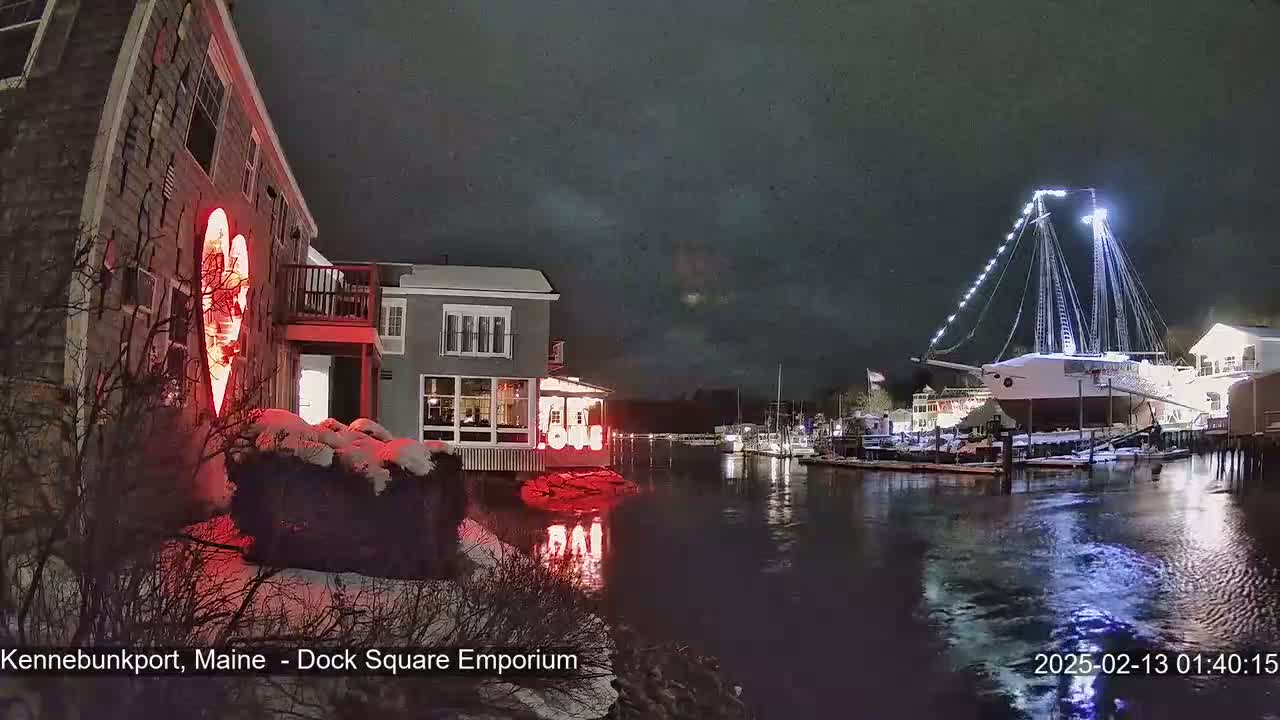 A nighttime scene of a snow-covered harbor with a large illuminated sailboat, buildings with lights, and a heart-shaped light display on one building.