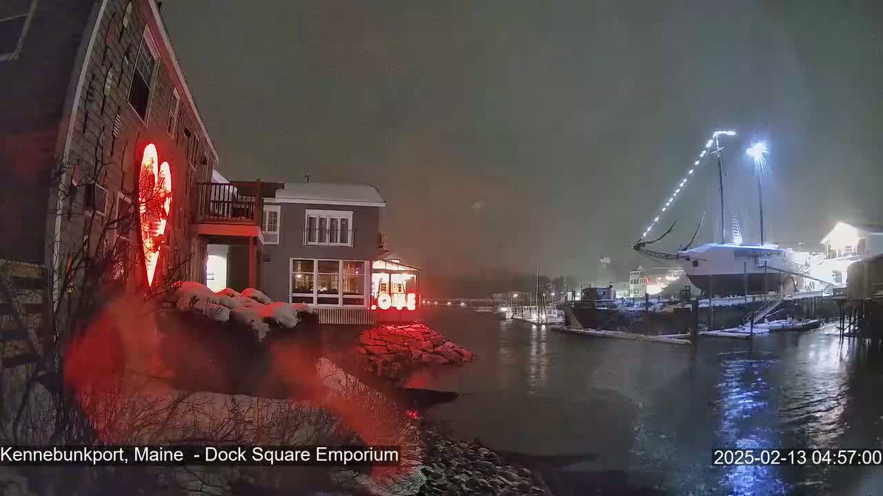 A snow-covered waterfront scene at night shows a building illuminated with a large red heart, a docked sailboat with lights, and other buildings across a calm waterway.