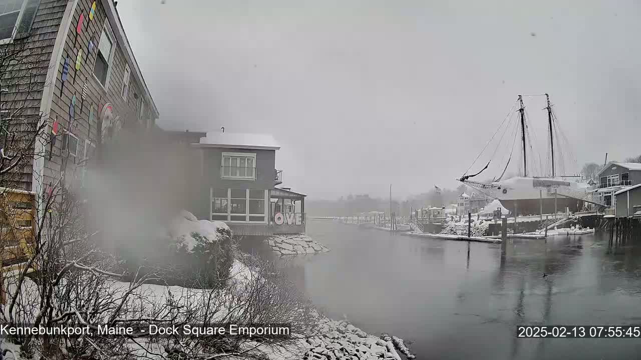 A snow-covered waterfront scene shows a docked sailboat and several buildings, with light snow falling.