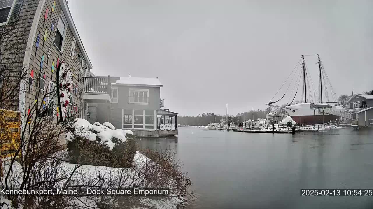 A snow-covered waterfront scene shows buildings and a large sailboat docked in a calm harbor under an overcast sky.