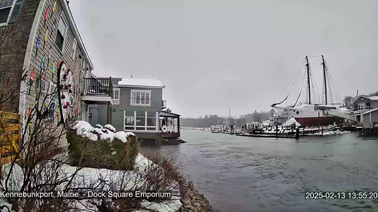 A snow-covered waterfront scene shows a canal with a large sailboat docked, smaller boats, and buildings along the snow-dusted banks.