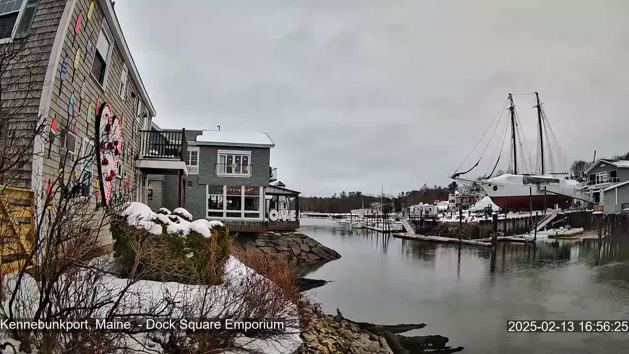 A snow-covered waterfront scene shows a building with colorful exterior art, other buildings, and a large sailboat docked in a calm harbor under an overcast sky.