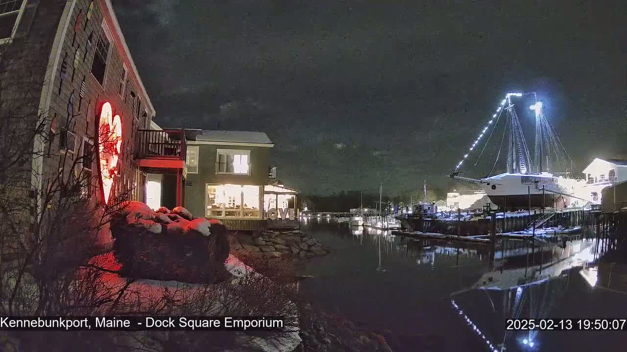 A snow-covered waterfront scene at night shows a building with a large illuminated heart on its side, another building with lit windows, and a large sailboat docked, all reflected in calm water.