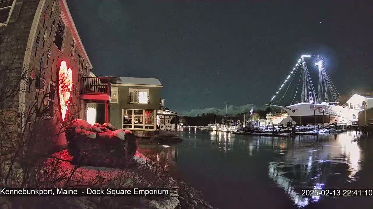 A nighttime harbor scene shows snow-covered ground, illuminated buildings, a large sailboat, and a red heart-shaped light on a building wall.
