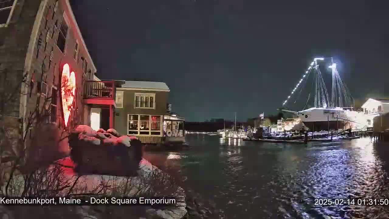 A nighttime waterfront scene shows a large sailing ship docked near illuminated buildings, a large red heart-shaped light on a building, and snow on the ground.