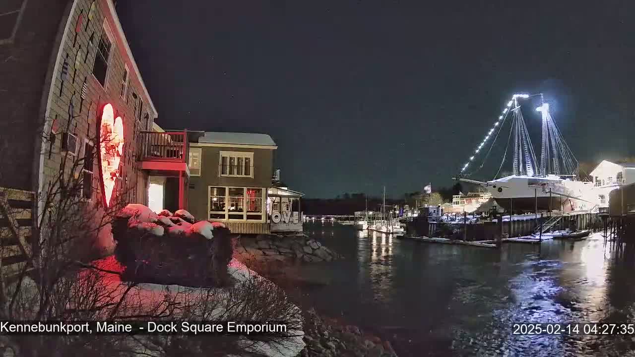 A nighttime harbor scene shows a large ship docked, illuminated buildings on the shore with a large red heart on one building, and snow-covered ground.