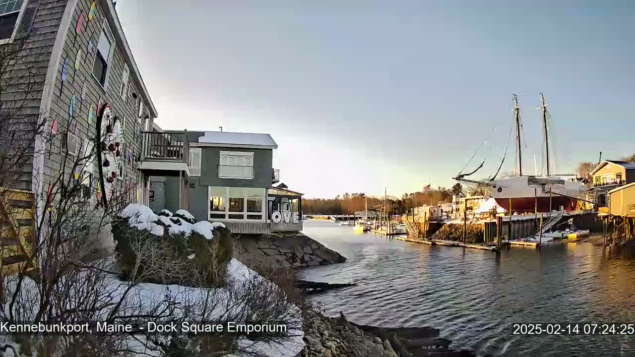 Snow-dusted buildings and a large sailboat are seen on a calm waterfront under a clear, bright sky.