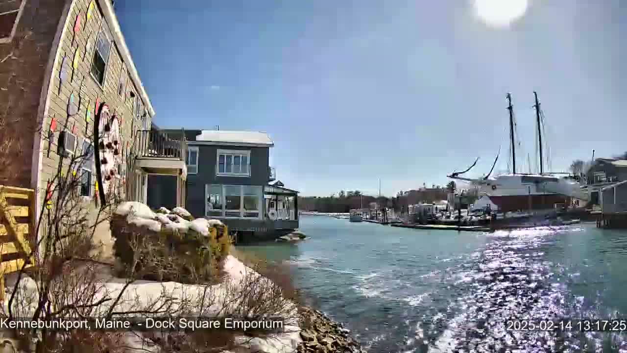 A colorful building with snow on the ground sits next to a calm waterway with a large sailboat docked in the background under a sunny sky.