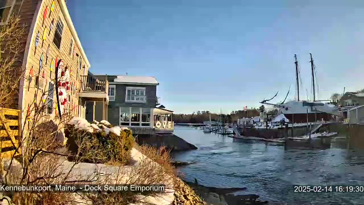 A waterfront scene on a sunny day shows a building adorned with colorful decorations next to a calm body of water with several boats and a large sailboat docked nearby.
