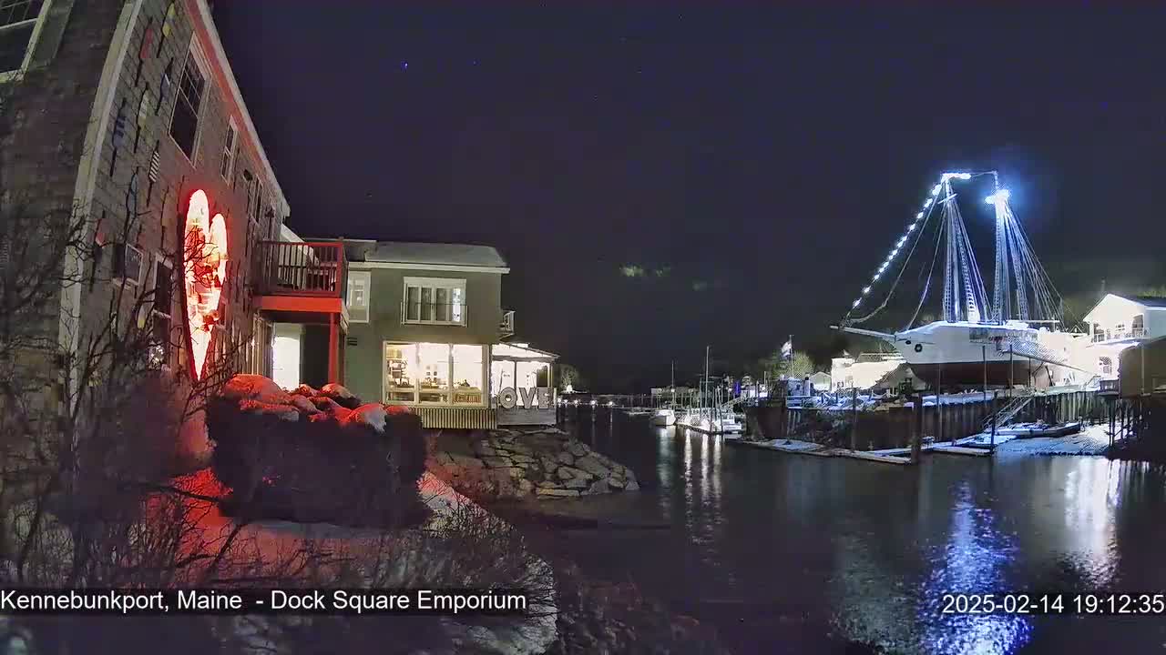 A nighttime scene of a snow-dusted harbor, featuring a large illuminated sailboat docked near a building with a large illuminated heart on its side.