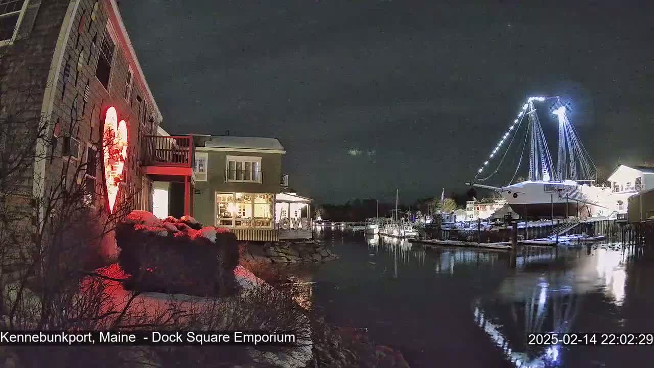 A nighttime scene shows a snow-covered waterfront with buildings illuminated, a large sailboat lit up at a dock, and a red heart-shaped light display on a building.