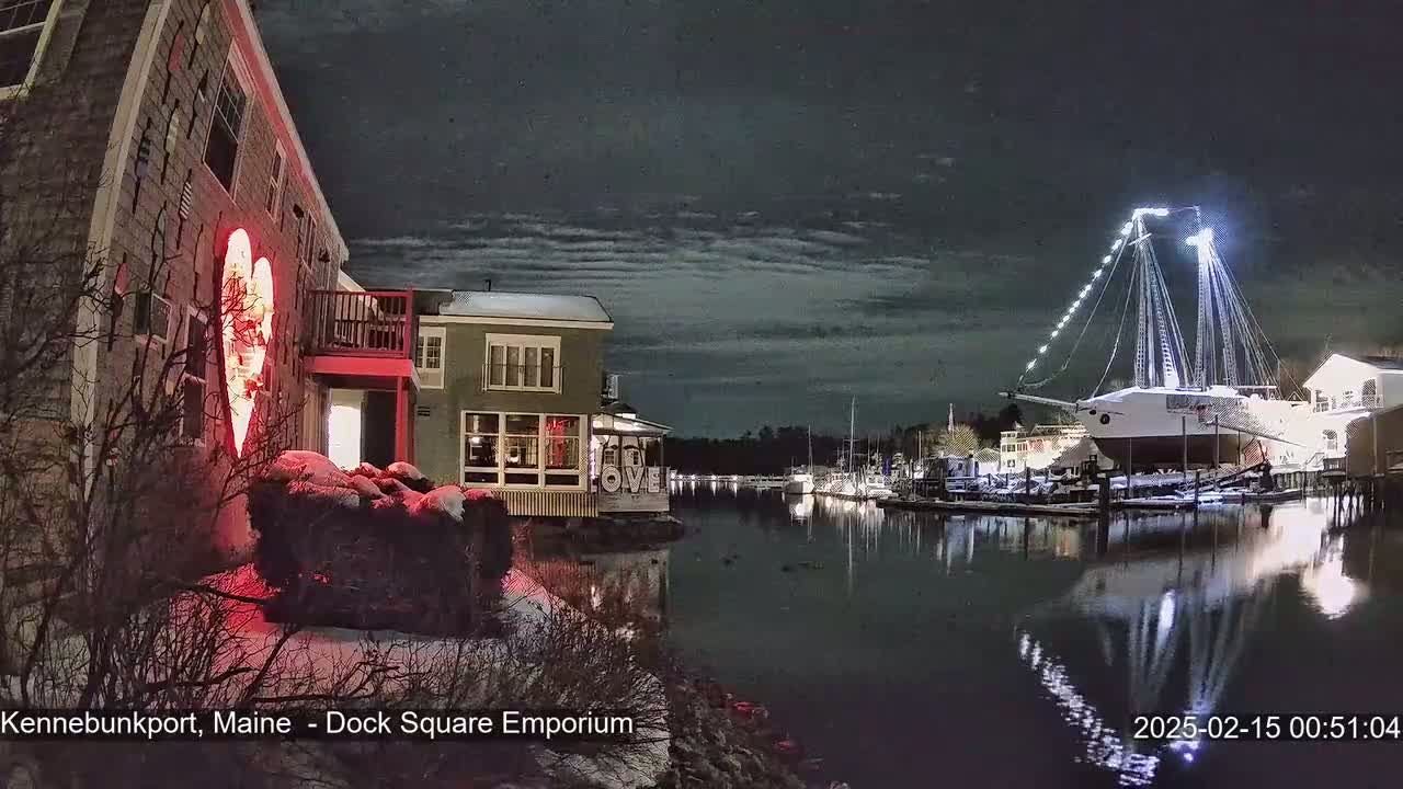 A snow-covered waterfront scene at night shows a large illuminated sailboat docked, buildings with lights on, and a large illuminated heart-shaped decoration on one building.