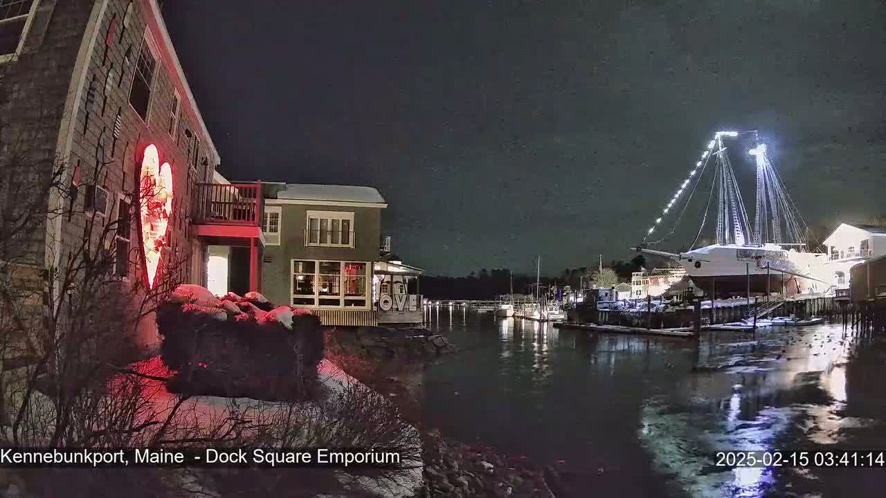 A nighttime scene of a harbor with a large sailboat illuminated, a building displaying a lighted heart, and snow on the ground.
