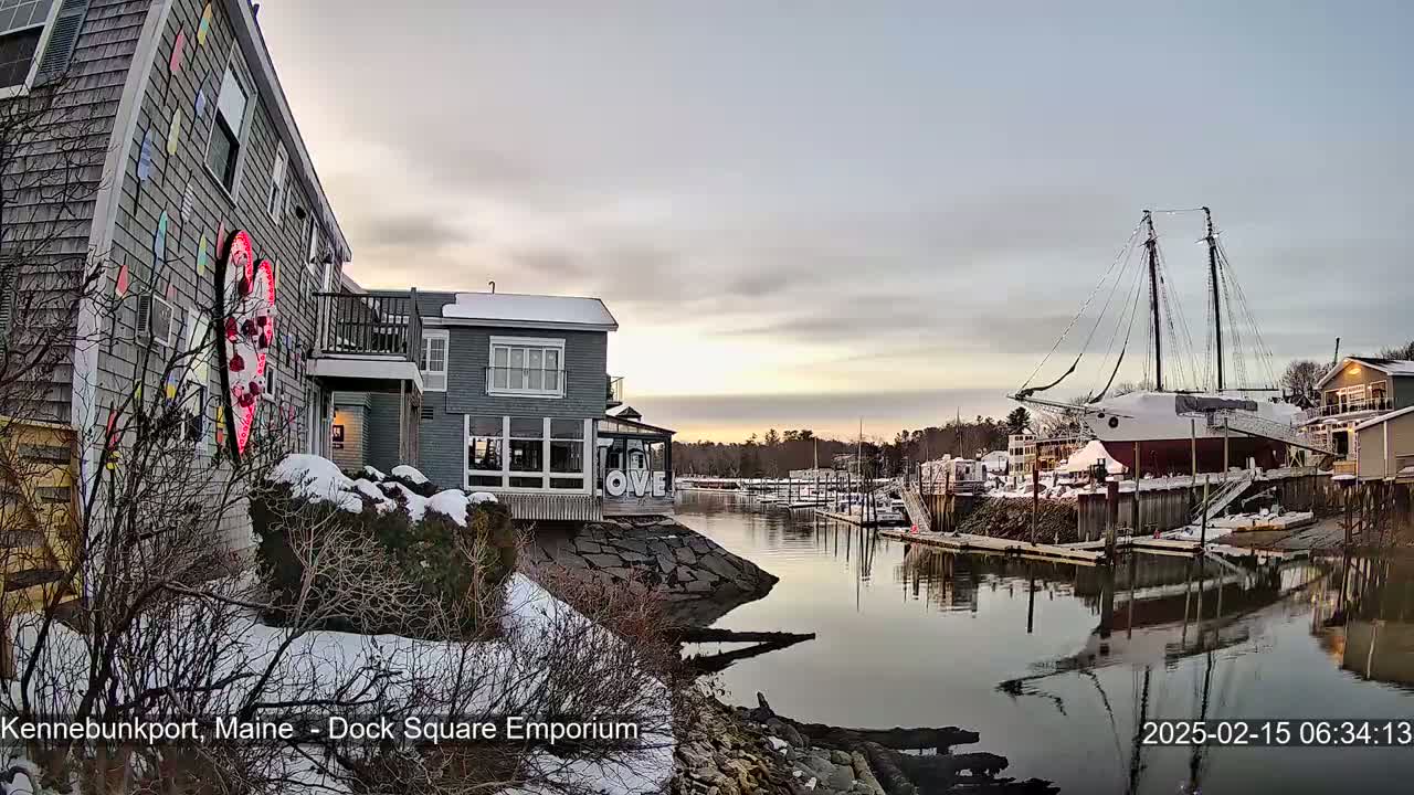 A snow-covered waterfront scene at sunset shows buildings, a docked sailboat, and calm water reflecting the sky and buildings.