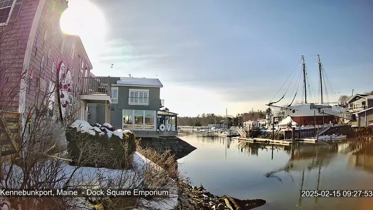A sunny winter day shows a waterfront scene with snow on the ground, buildings, and a large sailboat docked, all reflected in calm water.