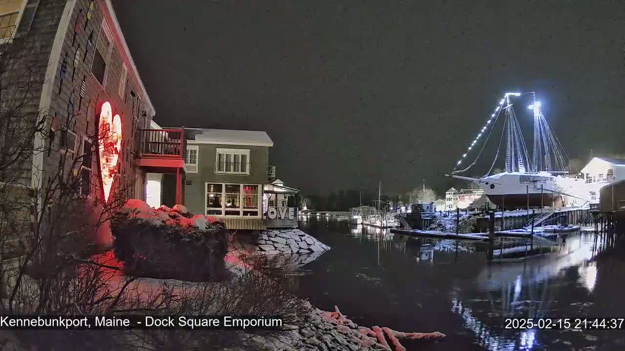 A snow-covered waterfront scene at night shows a building with a large illuminated heart on its side, other buildings, a docked ship illuminated with lights, and calm water reflecting the lights.