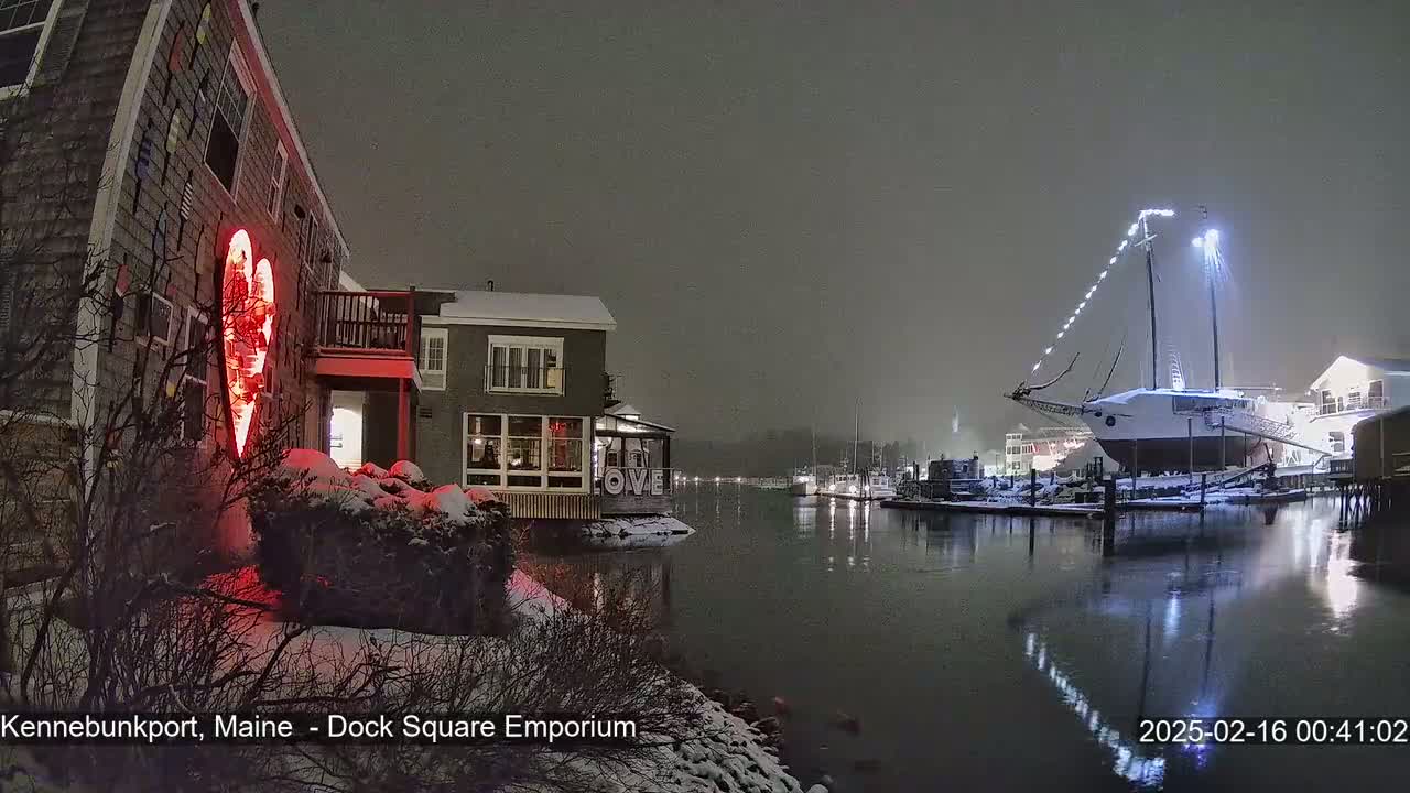 A snow-covered waterfront scene at night shows a building with a large red heart illuminated on its side, next to a calm harbor with a docked sailboat, all under a foggy sky.