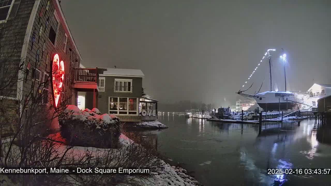 A snow-covered waterfront scene at night shows a building with a large illuminated red heart on its exterior, a docked sailboat with lights, and a calm waterway.