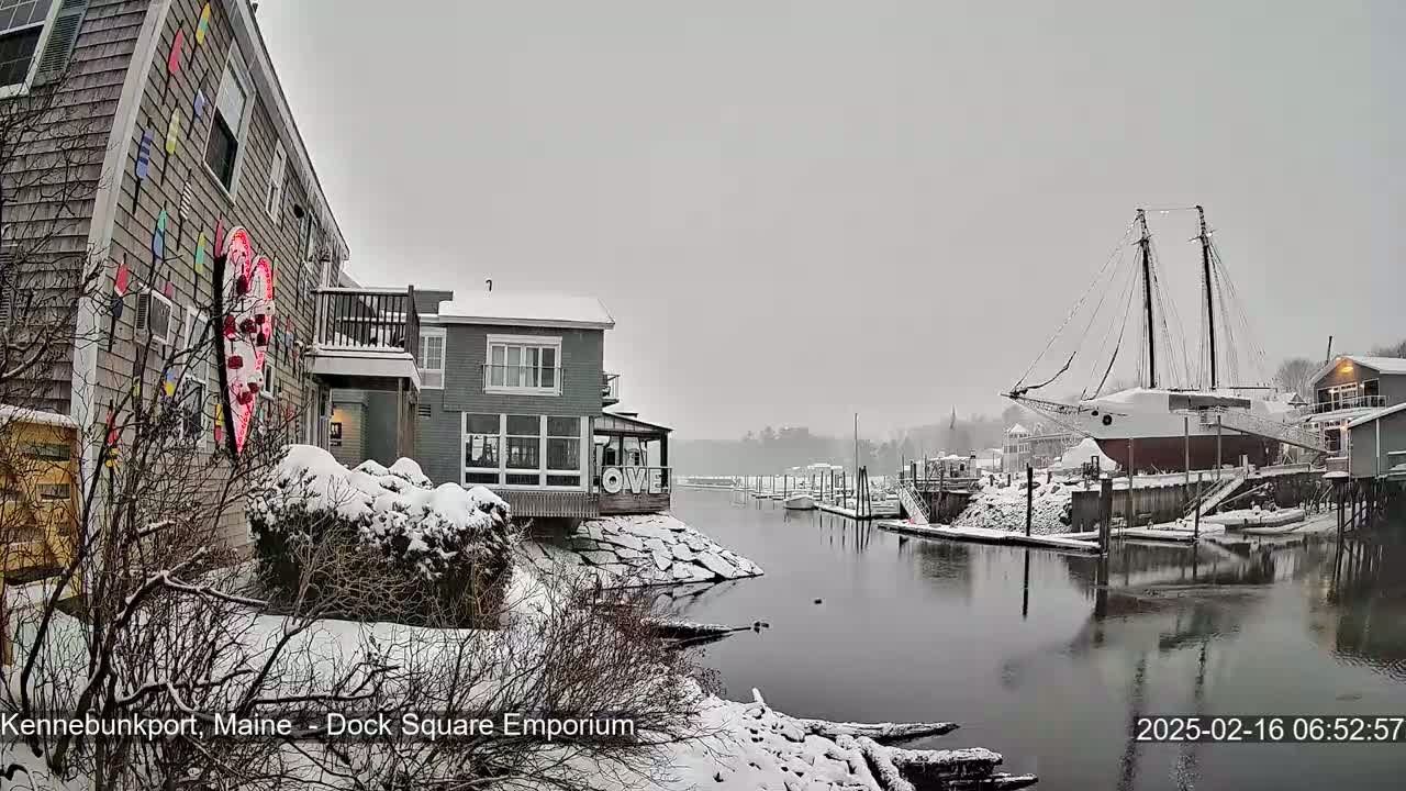 A snow-covered waterfront scene features buildings, a docked sailboat, and a calm waterway.