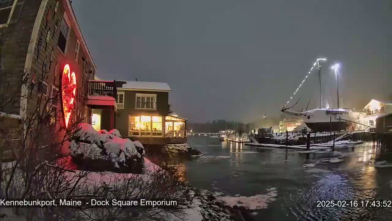 A snow-covered waterfront scene at night features a building with a large illuminated heart on its exterior, a docked sailboat, and partially frozen water.