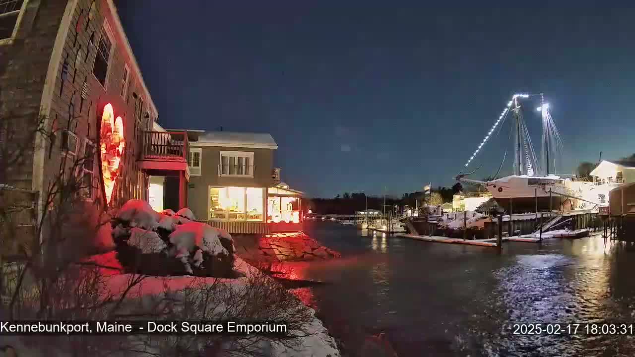 A snow-covered waterfront scene at night shows a building illuminated with a red heart-shaped light, other buildings with lit windows, and a large sailboat docked nearby under a clear, dark sky.