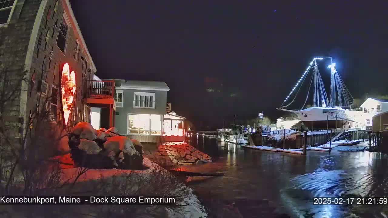 A snow-covered waterfront at night shows a building with a large illuminated heart on its side, other buildings, and a docked ship lit with white lights.