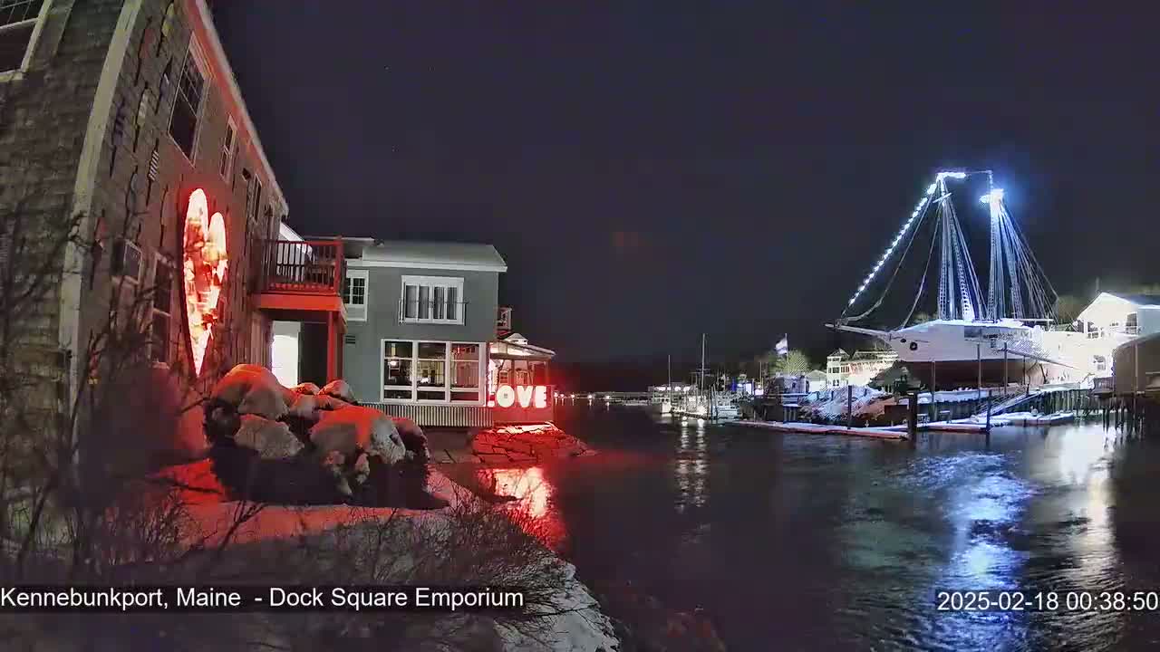 A nighttime waterfront scene shows a building with a lit-up heart and the word "LOVE," a large ship illuminated with lights, and snow-covered ground along the water's edge under a clear, dark sky.