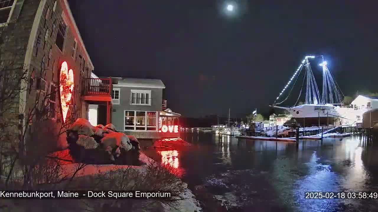 A nighttime waterfront scene shows a snow-covered building with a large illuminated heart, a "LOVE" sign, and a large ship docked nearby under a partly cloudy moonlit sky.