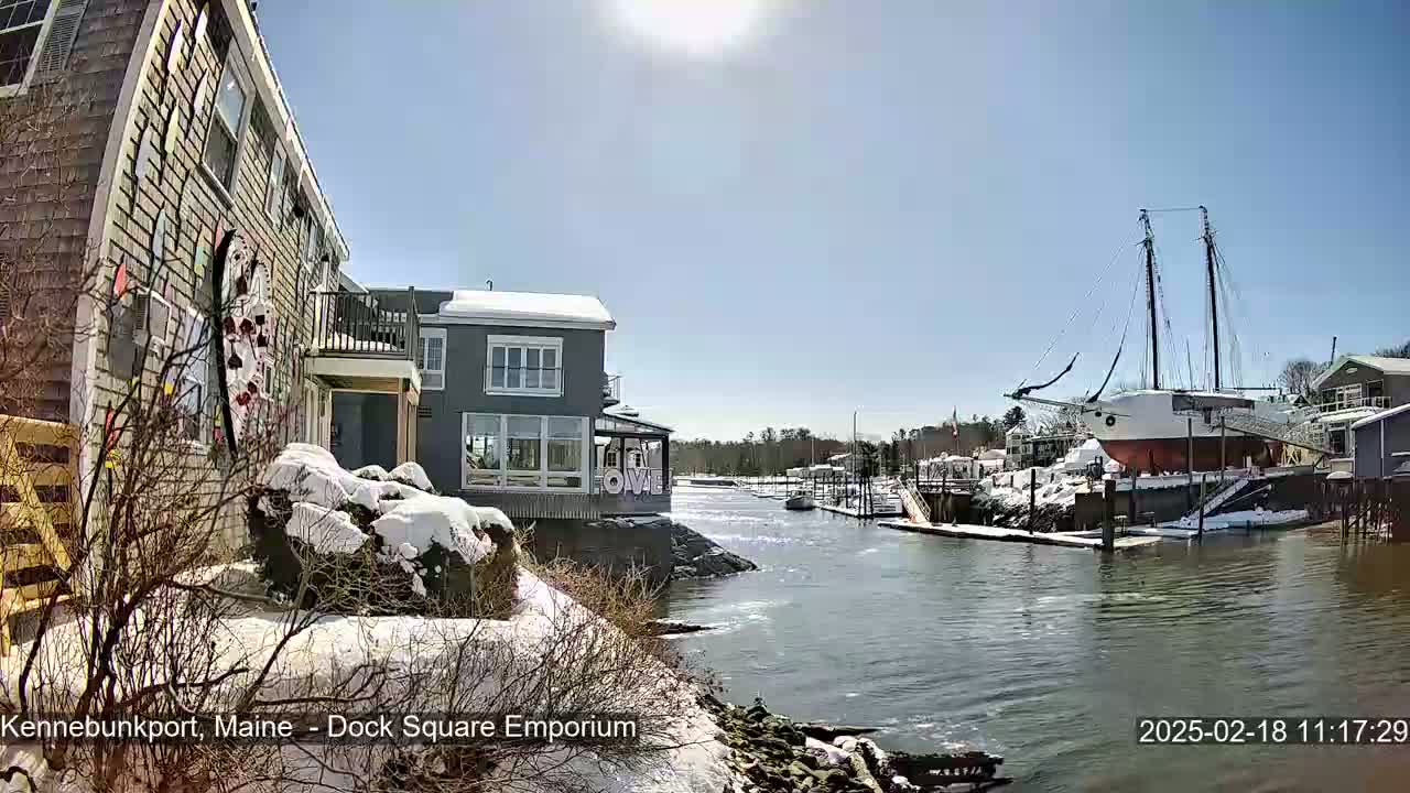 A sunny day shows a waterfront scene with snow on the ground and buildings, including a large sailing ship docked next to a small waterway.