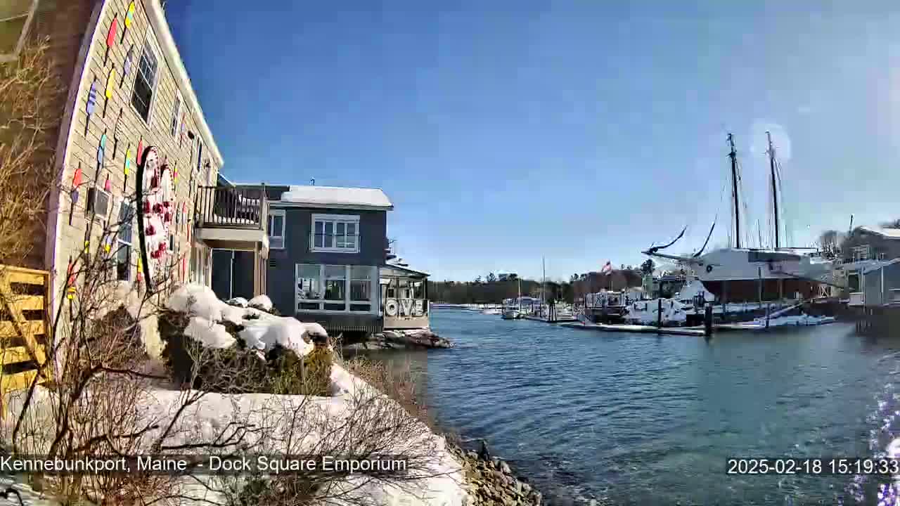 Snow-covered waterfront buildings and docked sailboats are visible on a sunny day.