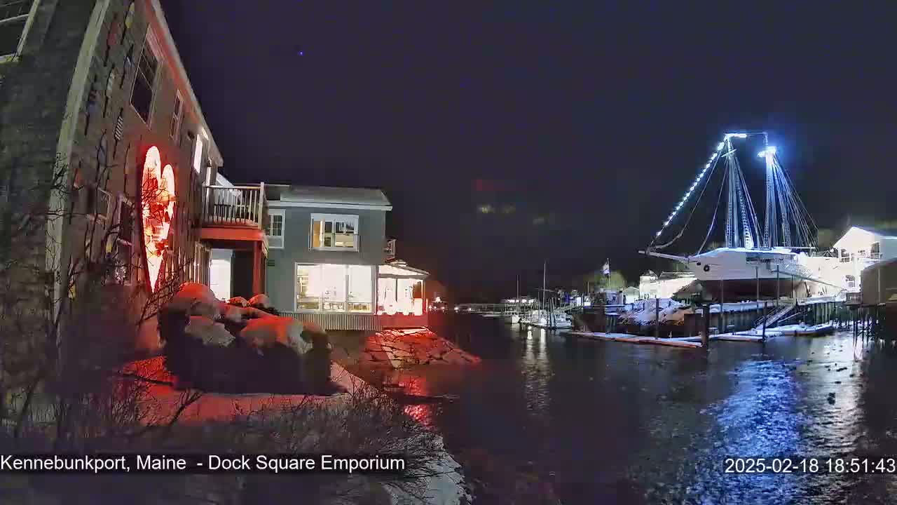 A snow-covered waterfront at night features a building with a large illuminated heart on its exterior, and a docked, illuminated sailing ship.