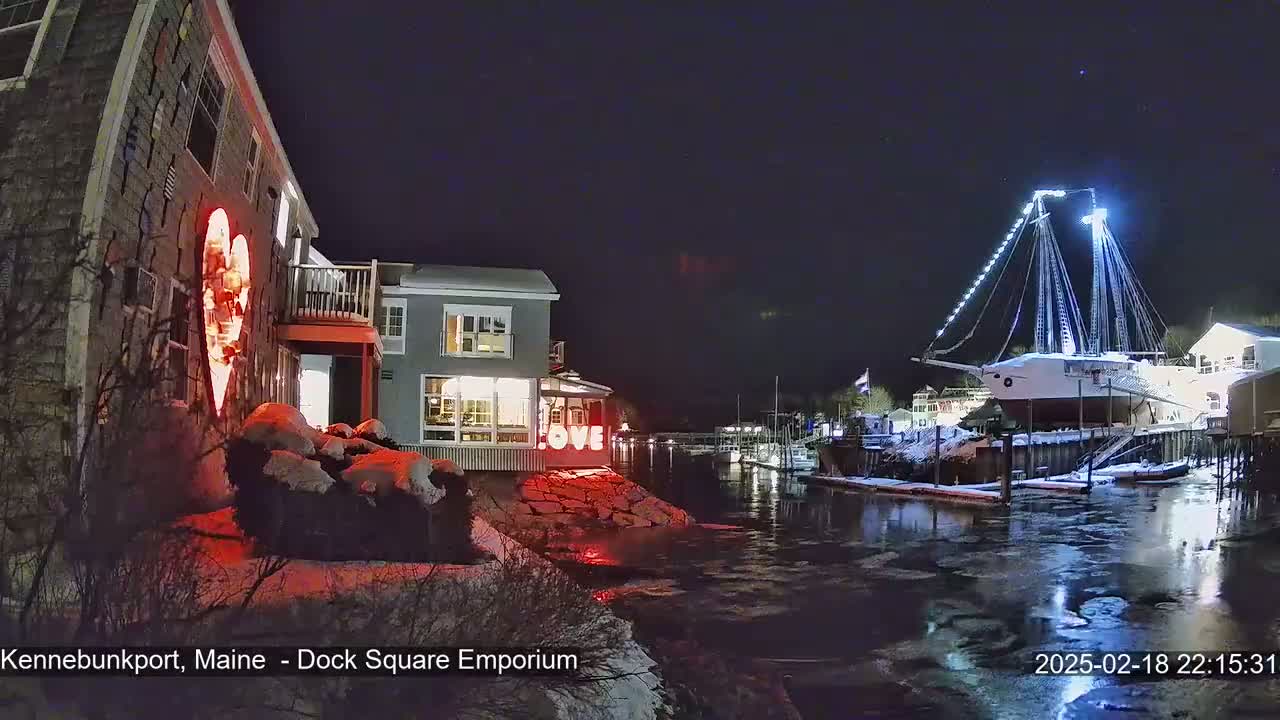 A nighttime scene shows a snow-dusted waterfront with illuminated buildings, a large sailboat, and a red heart-shaped light display.
