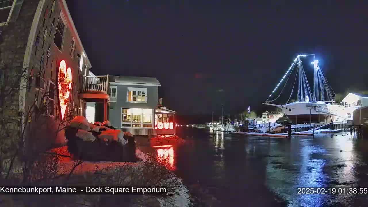 A snow-covered waterfront scene at night shows a building with illuminated heart and "LOVE" signs, a large illuminated ship, and other boats docked along a calm waterway.