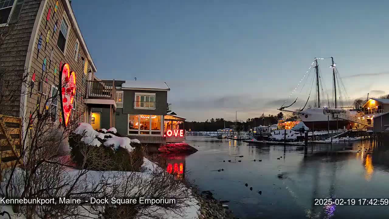 A snow-covered waterfront scene at twilight shows a building with a large illuminated heart and the word "LOVE" displayed, a docked sailboat with lights, and calm water reflecting the buildings and lights.
