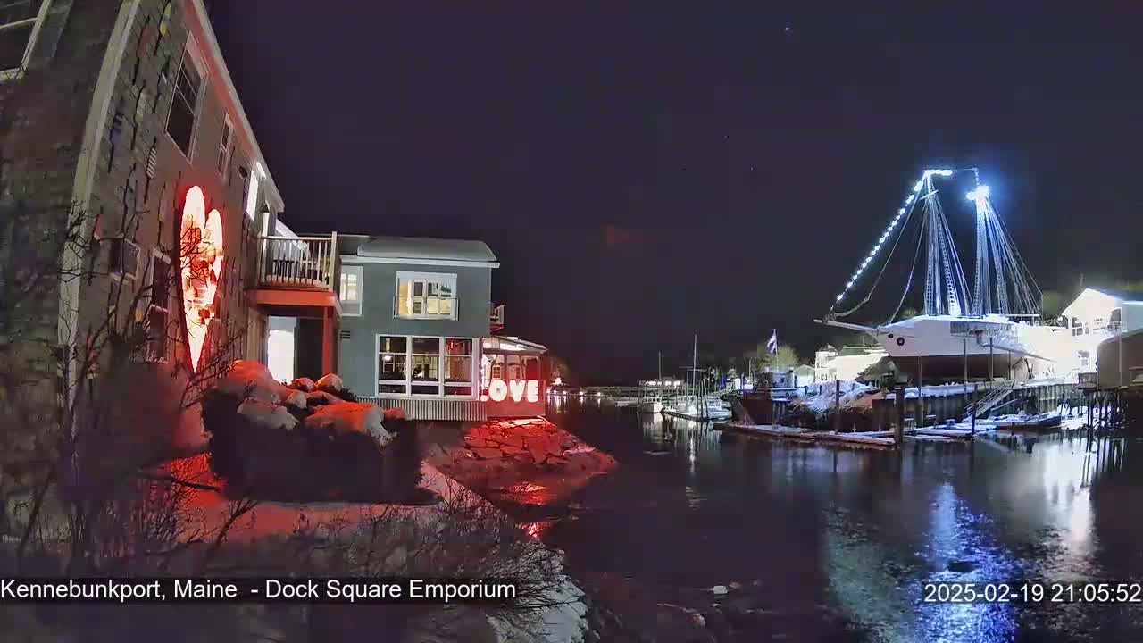 A nighttime waterfront scene shows a building with a large illuminated heart and the word "LOVE" displayed, alongside a docked, illuminated sailboat in a snow-dusted harbor.