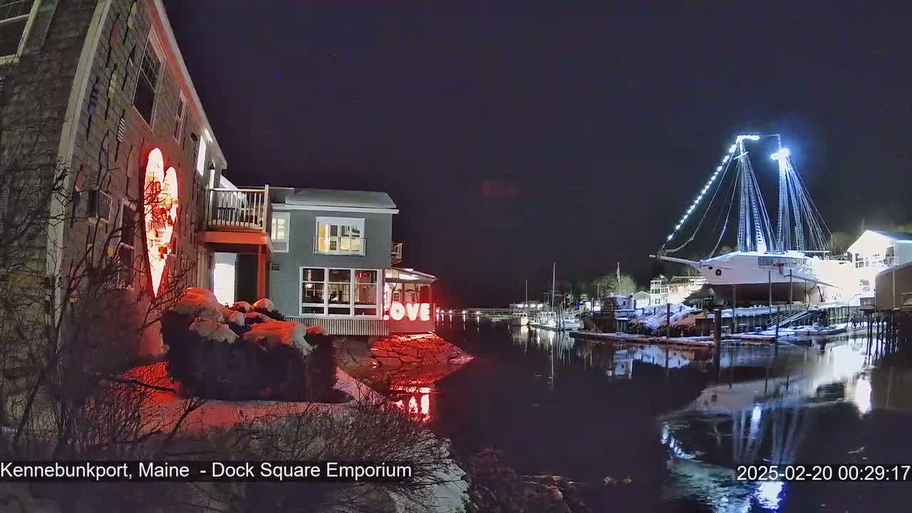 A nighttime waterfront scene shows a snow-dusted harbor with a large sailboat illuminated, buildings with illuminated hearts and signs, and calm water reflecting the lights.