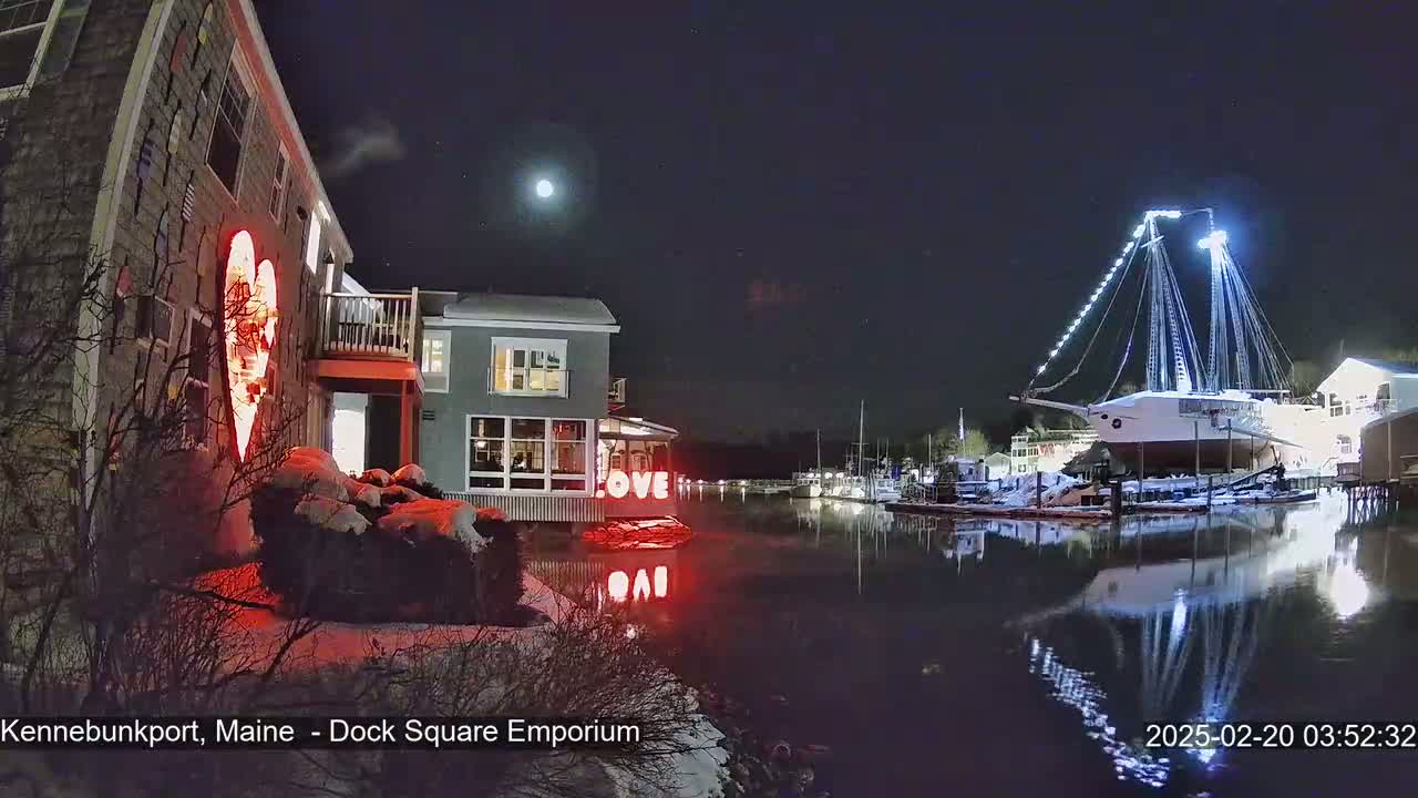 A snow-covered waterfront scene at night shows a building with a lit-up heart, a large illuminated ship in the harbor, and a bright moon in the sky.