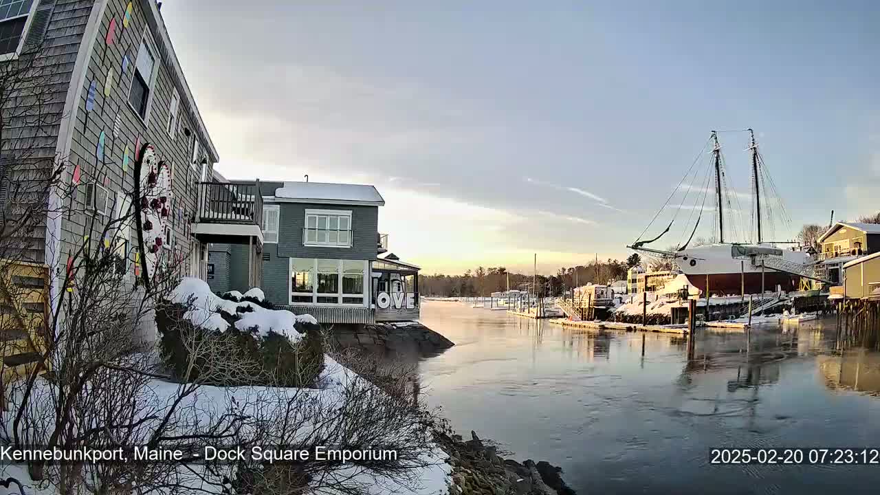 A snow-covered waterfront scene shows a canal with a large sailboat docked, and buildings with snow on the roofs and ground.