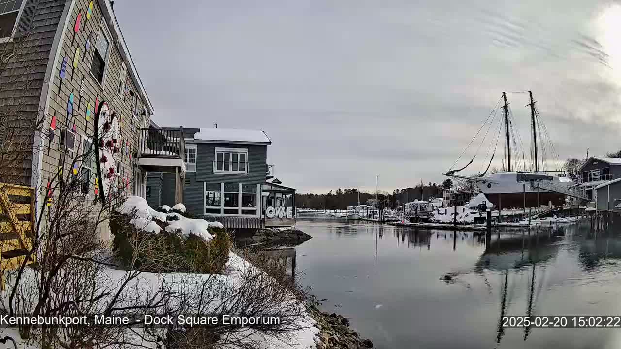 A snow-covered waterfront scene shows a calm waterway reflecting buildings and a large sailboat under an overcast sky.
