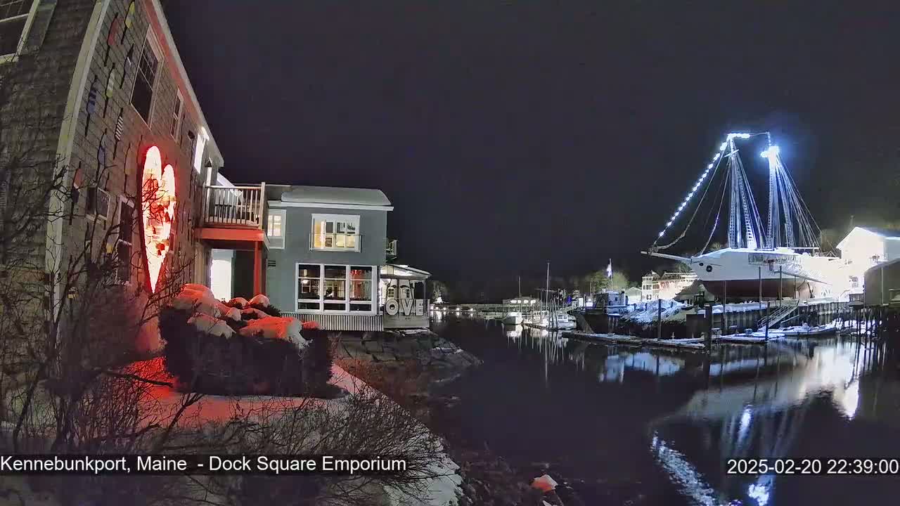 A nighttime view of a snow-covered waterfront scene with illuminated buildings, a large sailboat decorated with lights, and a red heart-shaped light on a building.