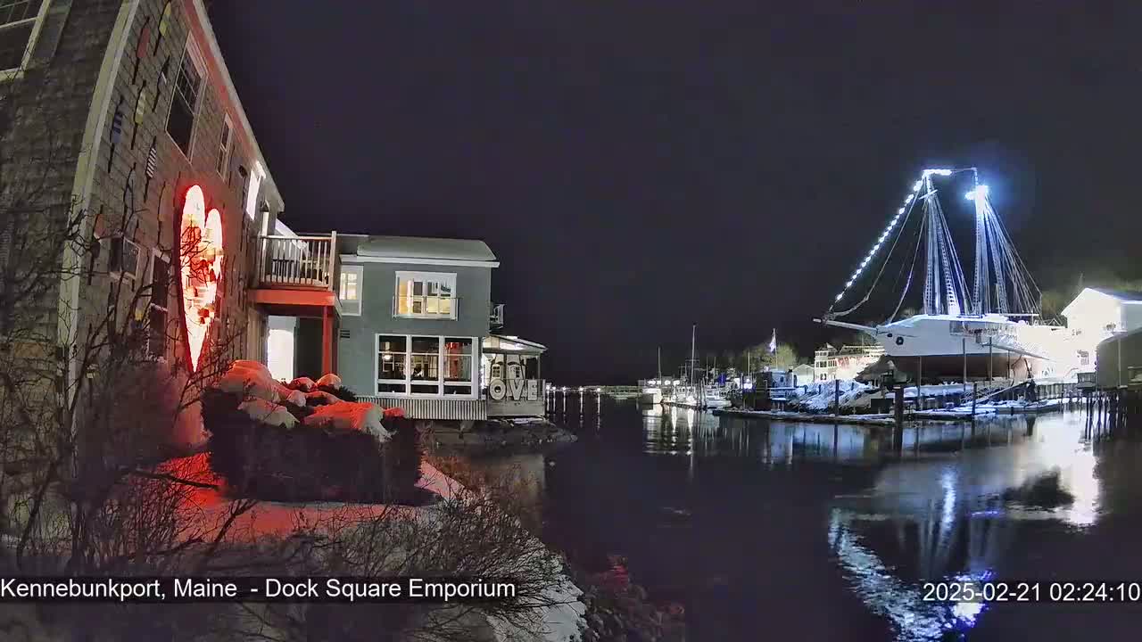 A nighttime waterfront scene shows a building with a large illuminated heart on its side, snow on the ground, and a large sailboat docked across the water.