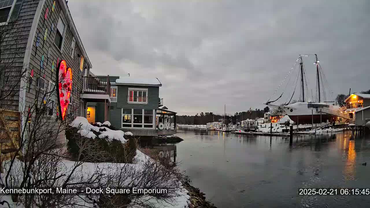 A snow-covered waterfront scene shows a building with a large illuminated heart on its side, another building across a calm waterway, and a docked ship under an overcast sky.