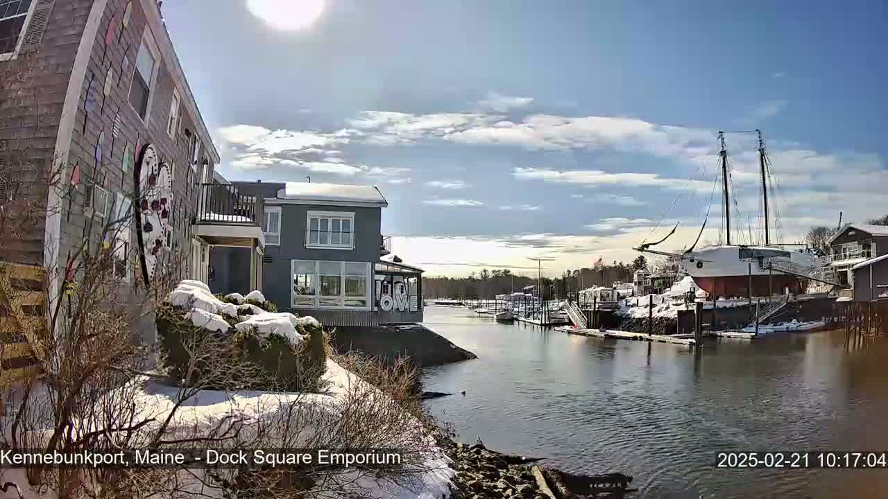 A partly sunny day with snow on the ground shows a harbor view with buildings, a large sailboat, and other small boats docked.